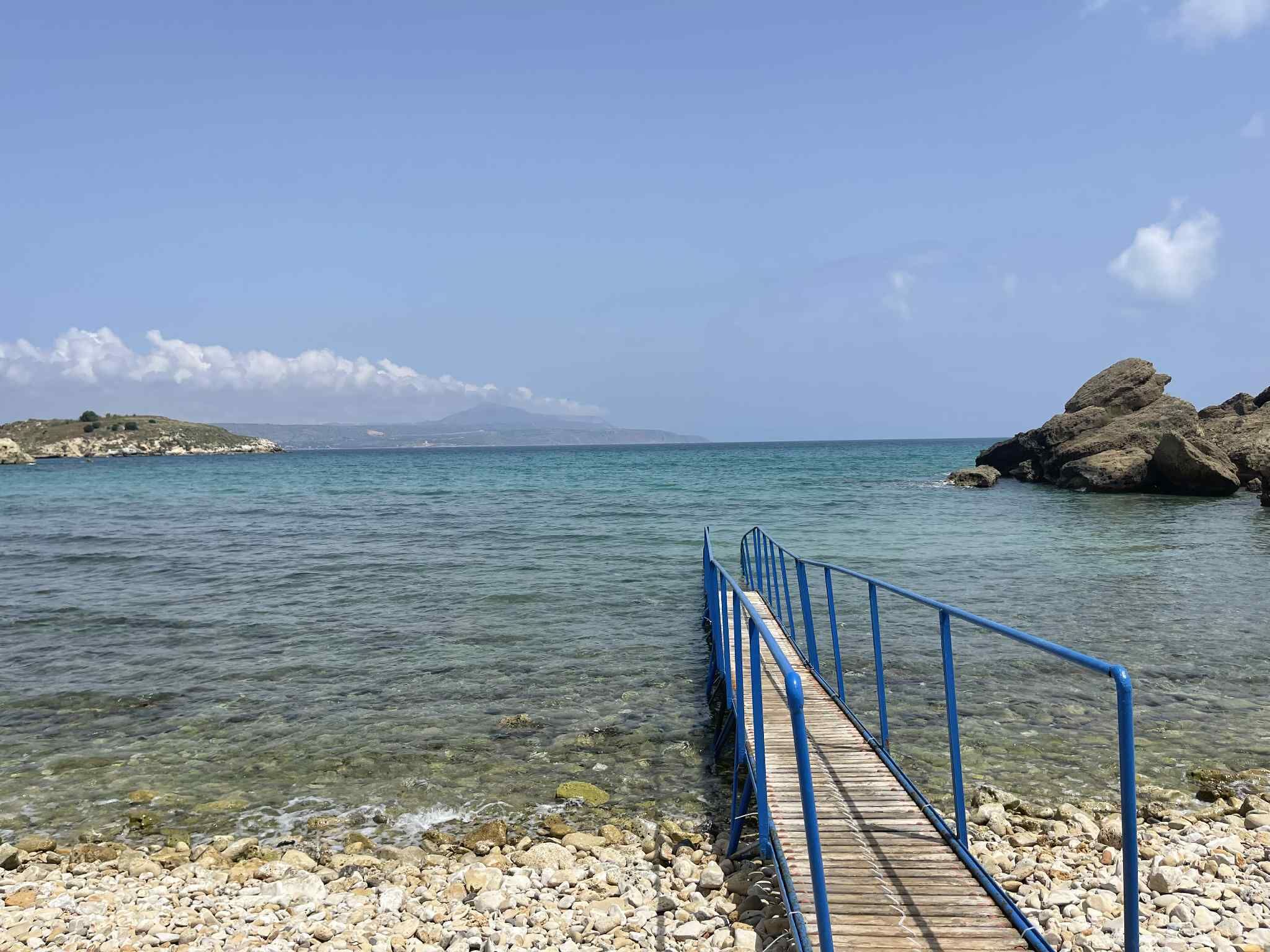 Cielo azul despejado, mar tranquilo, costa rocosa y muelle de madera con barandas azules.