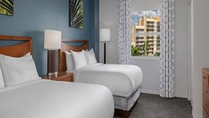 A hotel room with two beds, a nightstand, a lamp, a window with a view of buildings, and patterned curtains.