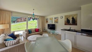 A modern living room with a sofa, a glass dining table, and a kitchen area with a TV and a sink. - Cornelia 11 Appartement/Fewo 6-Bettwohnung by Interhome (Saas-Fee)