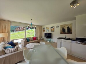 A modern living room with a sofa, a glass dining table, and a kitchen area with a TV and a sink. - Cornelia 11 Appartement/Fewo 6-Bettwohnung by Interhome (Saas-Fee)