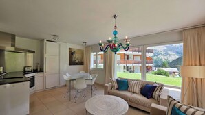 A modern living room with a sofa, dining table, and chairs, a kitchen area with a stove and cabinets, and a large window with a mountain view. - Cornelia 11 Appartement/Fewo 6-Bettwohnung by Interhome (Saas-Fee)