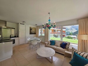 A modern living room with a sofa, dining table, and chairs, a kitchen area with a stove and cabinets, and a large window with a mountain view. - Cornelia 11 Appartement/Fewo 6-Bettwohnung by Interhome (Saas-Fee)