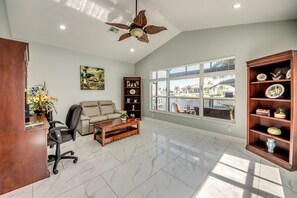 Uma sala de estar espaçosa com ventilador de teto, uma janela ampla com vista para um corpo d'água, um sofá, uma mesa de centro e uma estante de livros.