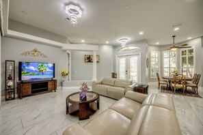 Ein großzügiges Wohnzimmer mit einem großen Flachbildfernseher, einem beigefarbenen Ledersofa, einem Couchtisch und einem Essbereich mit Tisch und Stühlen.