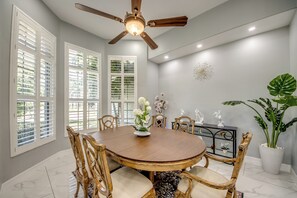 Uma sala de jantar com mesa redonda, cadeiras de madeira, ventilador de teto e um vaso com planta.