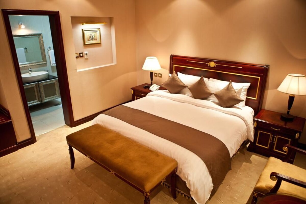 A hotel room with a large bed, a bench, a nightstand with a lamp, and a bathroom visible through an open door.