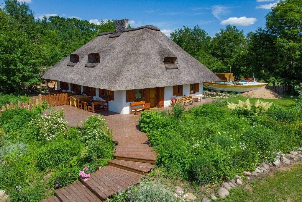 Une maison à toit de chaume, dotée d’une terrasse en bois menant à l’entrée, et entourée d’une végétation luxuriante.