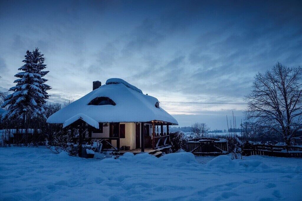Ett snötäckt hus med ett halmtak, omgivet av snötäckta träd och en klar himmel.