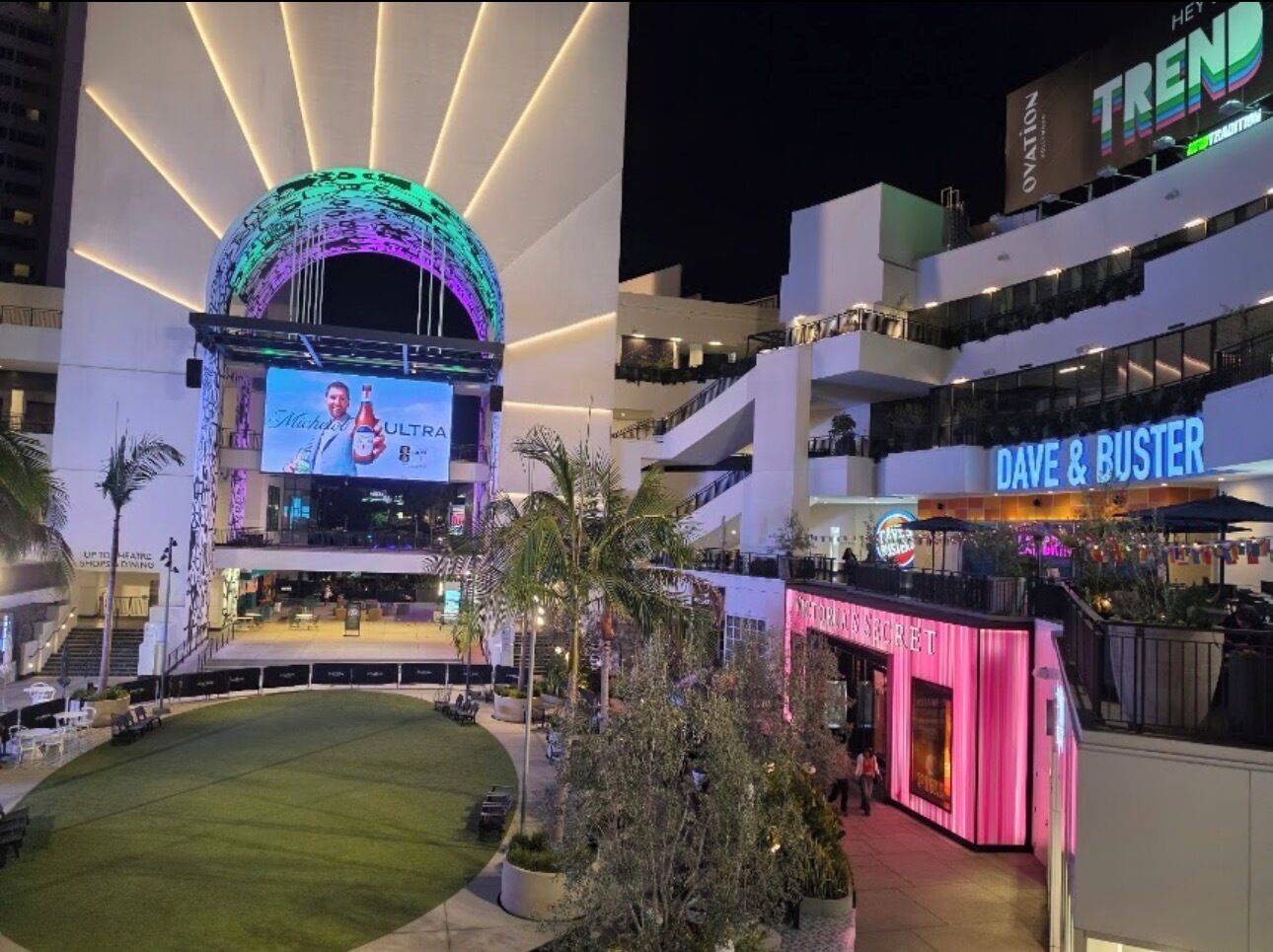 A modern building with a large digital screen displaying advertisements, a rooftop area with seating, and a brightly lit restaurant named 'Dave & Buster's.'