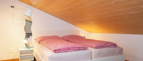 A bedroom with a sloped ceiling, a bed with checkered bedding, a bedside table with a lamp, and a wall-mounted light fixture.