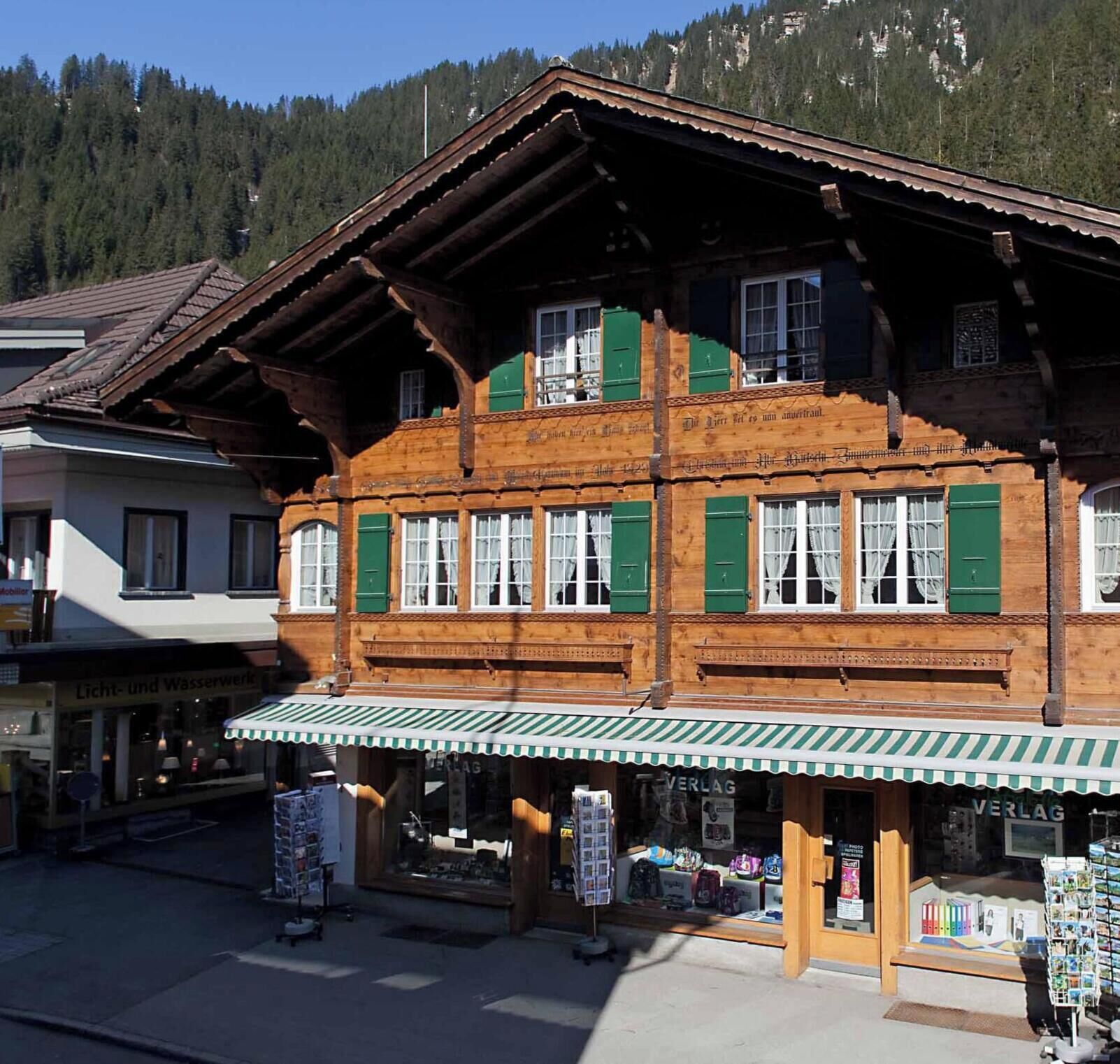 A traditional wooden building with a green awning, a visible shop window, and a mountain backdrop.