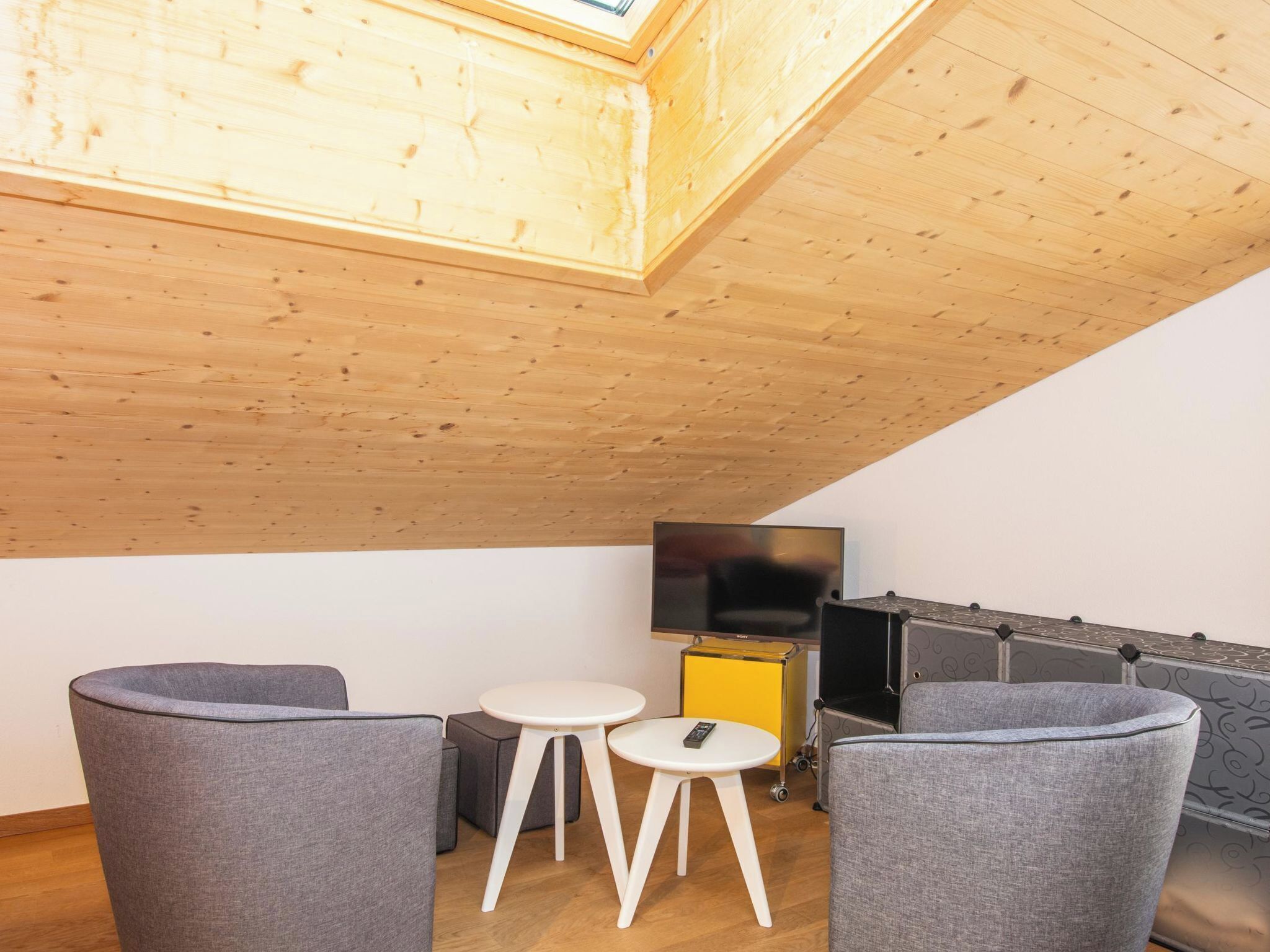 A modern living room with a wooden ceiling, a flat-screen TV, grey upholstered chairs, and a small white table.
