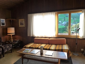 A cozy room with a bed, a table with magazines, a lamp, and a window with a view of a village.