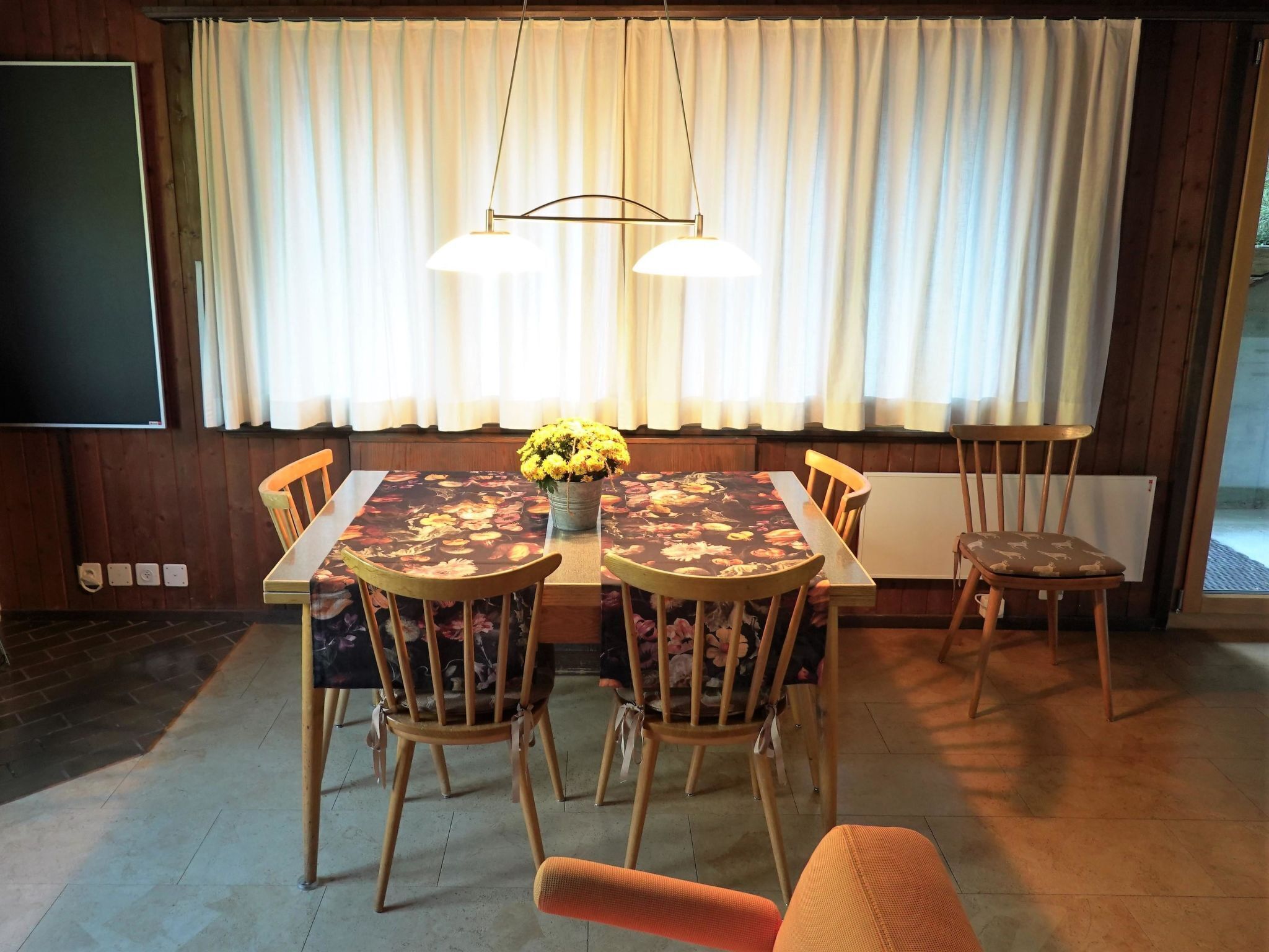 A dining area with a table, chairs, a vase with flowers, and a large window with curtains.