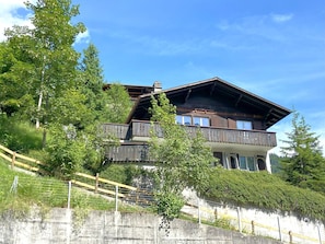 A two-story wooden chalet with a balcony, surrounded by lush greenery and a clear sky.