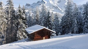 Eine Holzhütte mit schneebedecktem Dach, umgeben von schneebedeckten Bäumen und Bergen im Hintergrund.