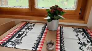 Ein Esstisch mit kariertem Tischläufer, eine Vase mit roten Blumen und ein Fenster mit Blick auf ein grünes Feld und Tiere.