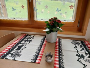 Ein Esstisch mit kariertem Tischläufer, eine Vase mit roten Blumen und ein Fenster mit Blick auf ein grünes Feld und Tiere.