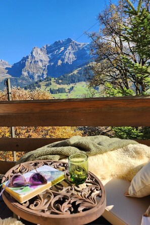 A wooden bench with a view of mountains, a book, a glass of drink, and a knitted blanket. - Juwel Appartement/Fewo, Dusche, WC, 2 Schlafräume 3-Zimmerwohnung by Interhome (Adelboden)