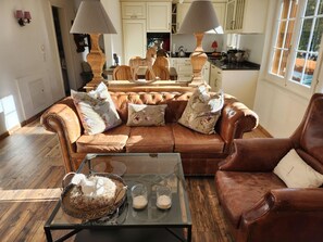 Ein Wohnzimmer mit braunem Ledersofa, einem Glastisch und einer Küche im Hintergrund.