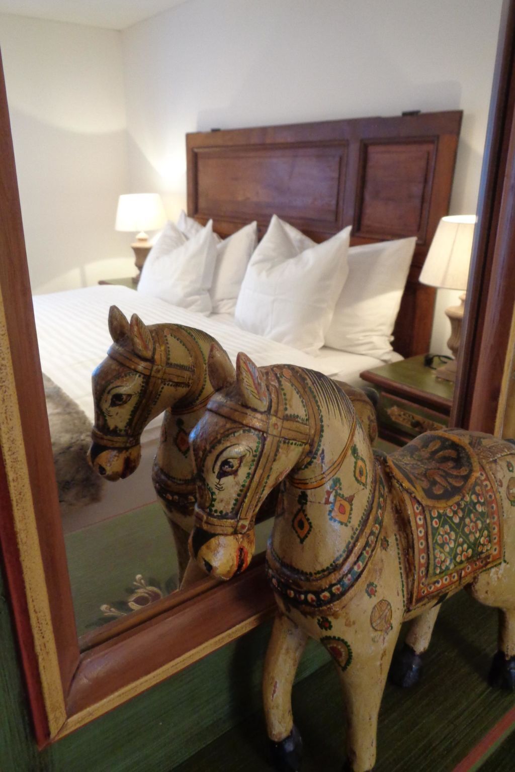 Een slaapkamer met een houten hoofdeinde, wit beddengoed en een spiegel die een decoratief paard weerspiegelt.