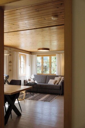 A room with a wooden ceiling, a grey sofa, a wooden dining table, and a window with curtains. - Gästehaus Alpengruss Appartement/Fewo, Bad, WC, 1 Schlafraum by Interhome (Adelboden)