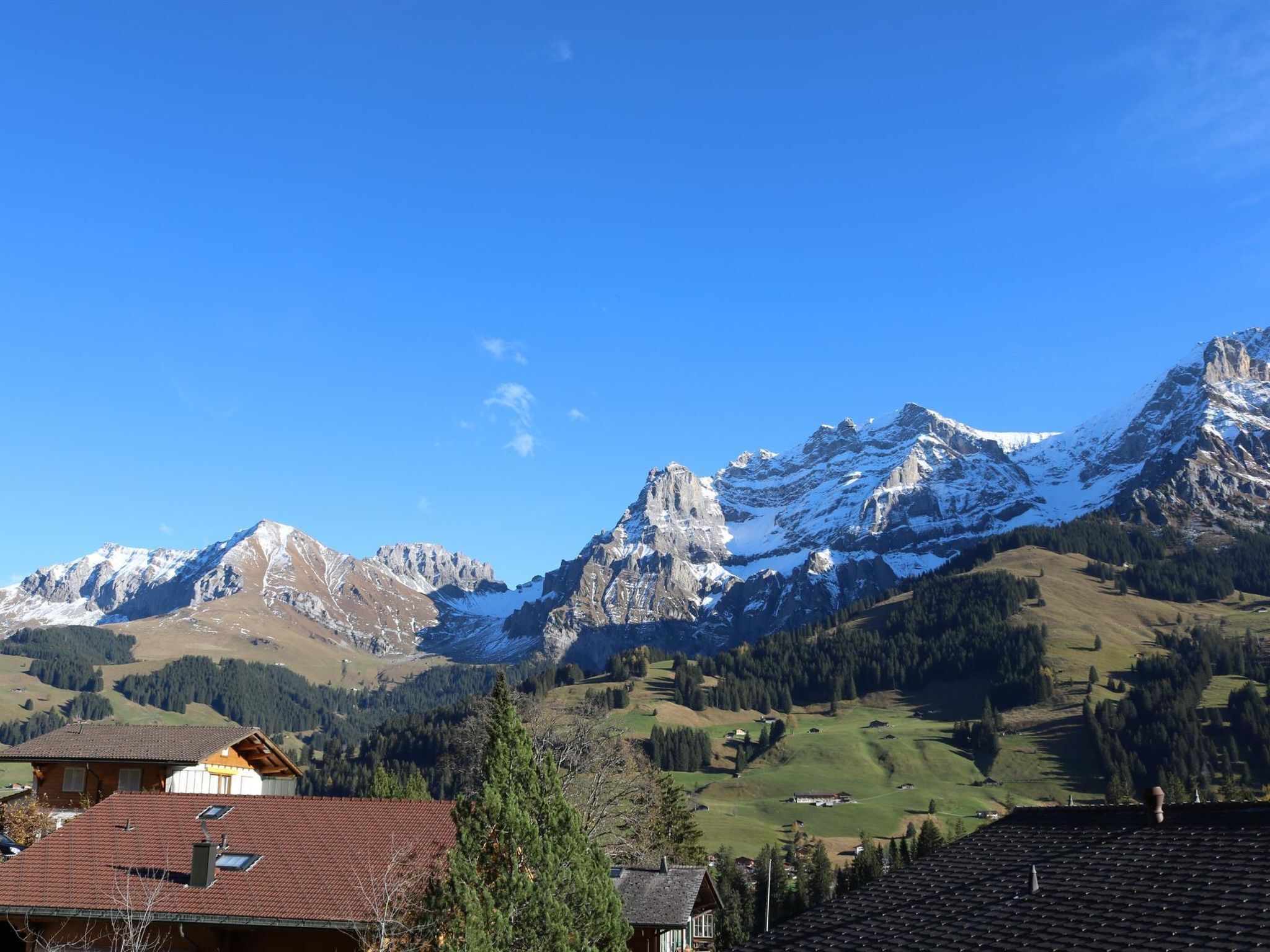 Paisaje montañoso con picos nevados, cielo azul despejado y un valle con casas y vegetación.