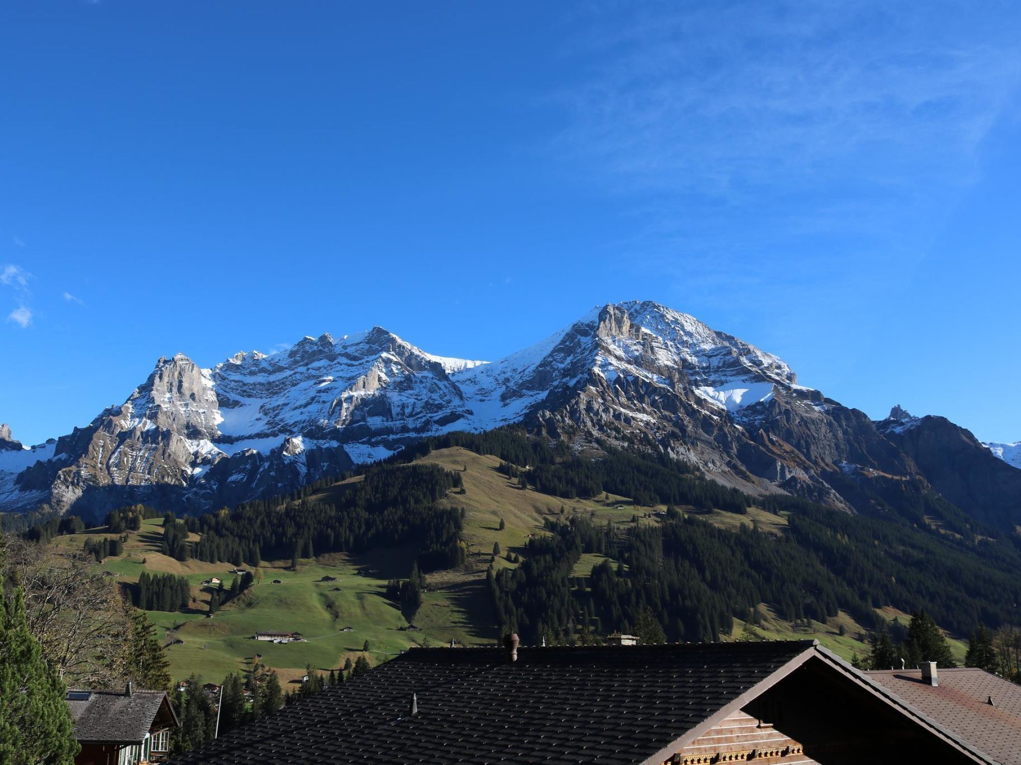 A mountain landscape with snow-capped peaks, a clear blue sky, and a view of a residential area with houses and a roof.