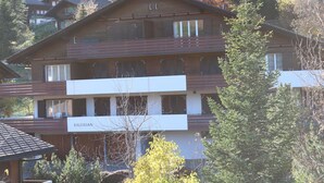 Ein mehrgeschossiges Gebäude mit einem Balkon im zweiten Stock, umgeben von Bäumen und anderen Gebäuden.