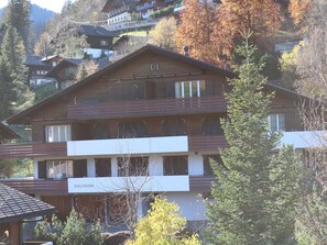 A multi-story building with a balcony on the second floor, surrounded by trees and other buildings. - Gruebi - Haus Baldrian Appartement/Fewo, Bad, WC, 2 Schlafräume 3-Zimmerwohnung by Interhome (Adelboden)