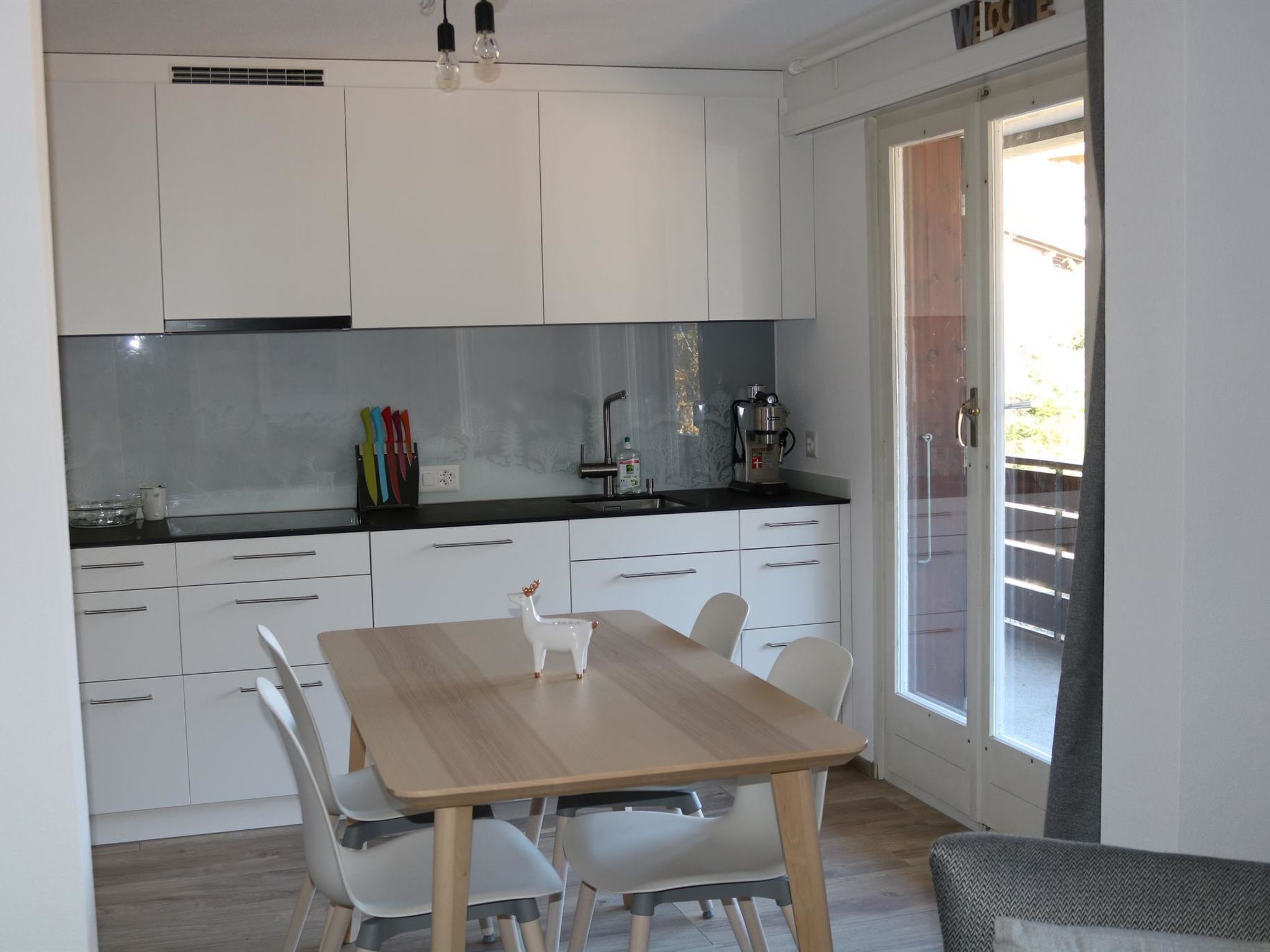 Una cocina moderna con armarios blancos, una mesa de comedor de madera y un ventanal que da a un balcón.