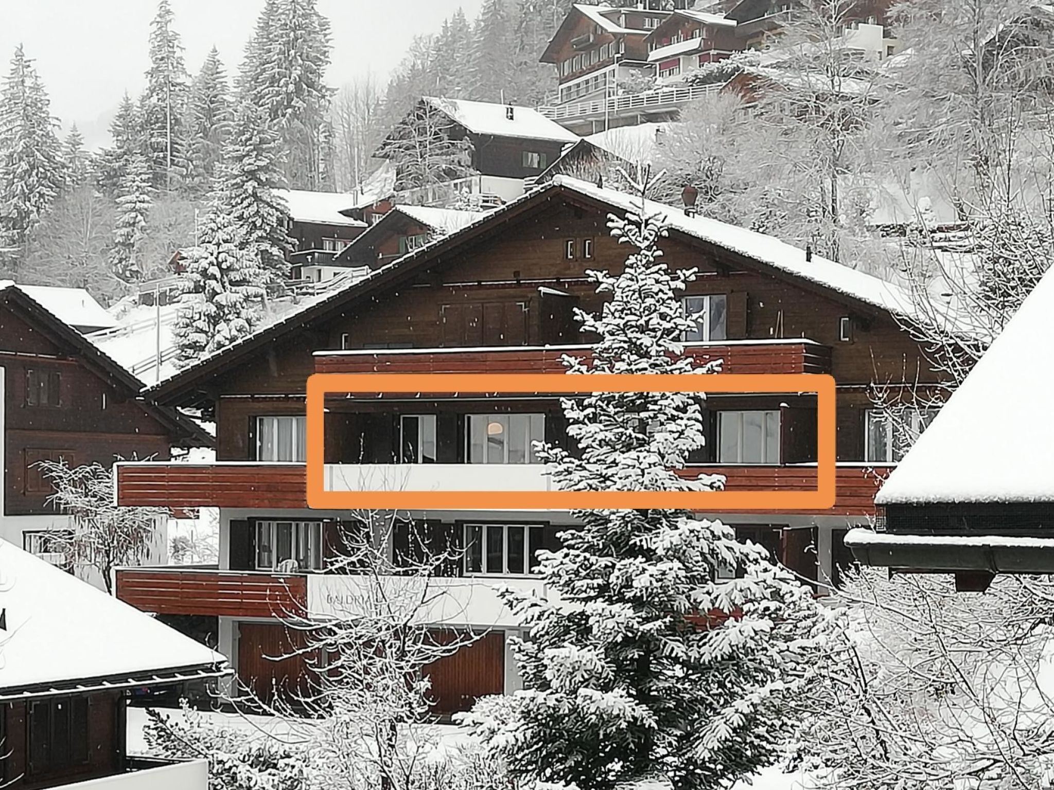 Een houten chalet met balkon, omringd door met sneeuw bedekte bomen en andere gebouwen.