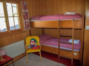 A bunk bed room with a red and white checkered bedspread, a yellow chair with a clown figure, and a window with a view of a house. - Chalet Sturmfang Appartement/Fewo, Dusche und Bad, WC, 4 oder mehr Schlafräume by Interhome (Adelboden)