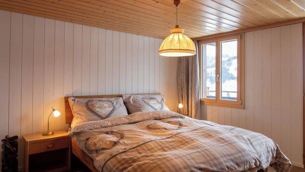 A bedroom with a wooden ceiling, a bed with a patterned bedspread, two bedside lamps, and a window with a view of the outdoors. - Waldrand Appartement/Fewo, Bad, WC, 2 Schlafräume 3-Zimmerwohnung by Interhome (Adelboden)