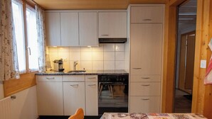 A compact kitchen with a built-in oven, a dining table with a patterned tablecloth, and a window with curtains. - Waldrand Appartement/Fewo, Bad, WC, 2 Schlafräume 3-Zimmerwohnung by Interhome (Adelboden)