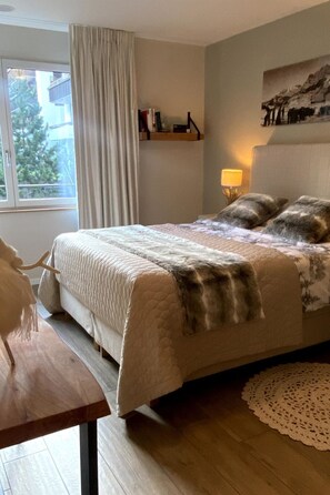 A bedroom with a bed, a bedside table, a lamp, a window with curtains, and a wall-mounted shelf with books. - Alpenrose Appartement/Fewo, Bad, WC, 3 Schlafräume 4-Zimmerwohnung by Interhome (Adelboden)