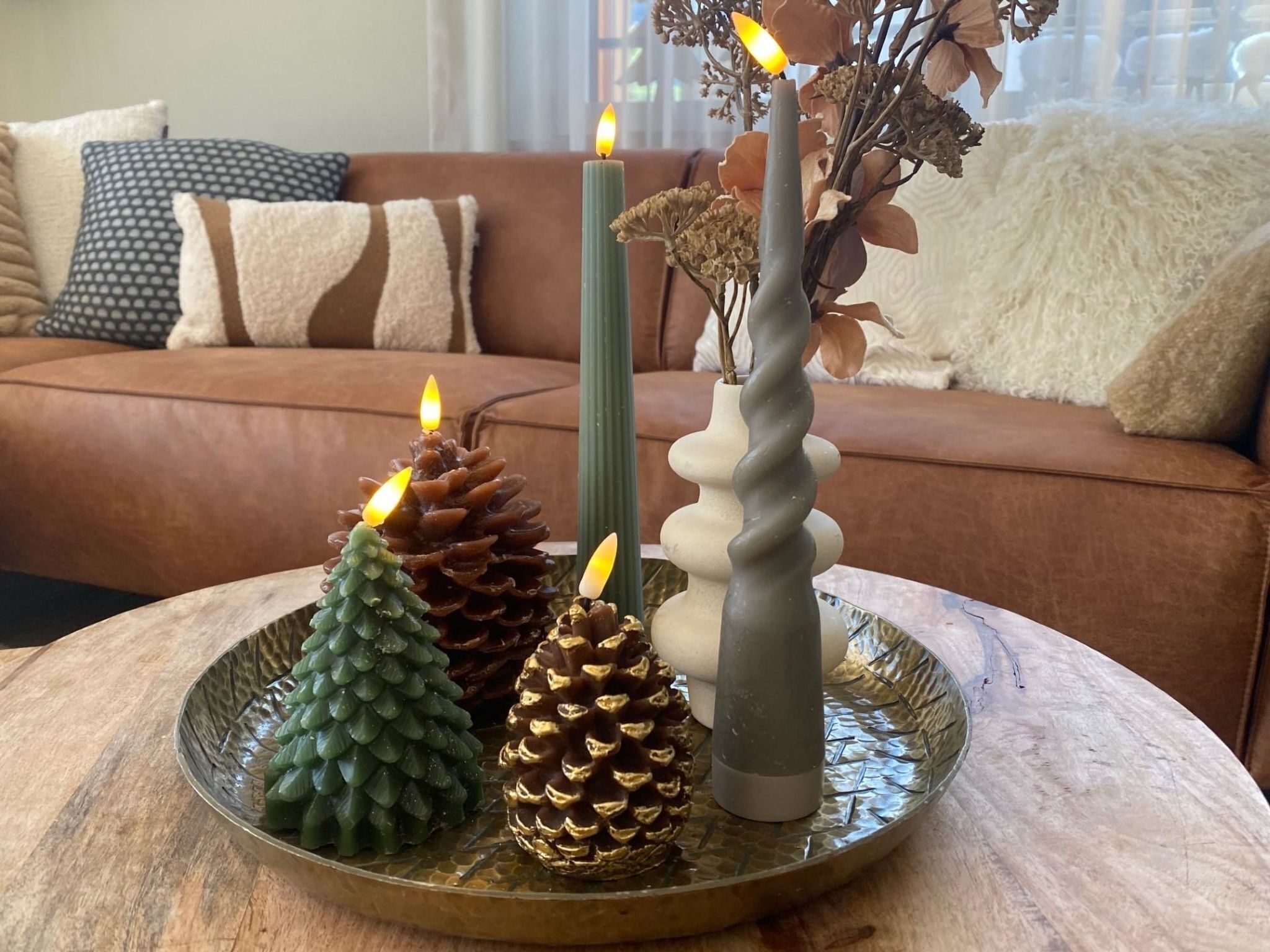 Una mesa redonda de madera con una bandeja decorativa que sostiene tres velas, una piña y un florero con flores secas.
