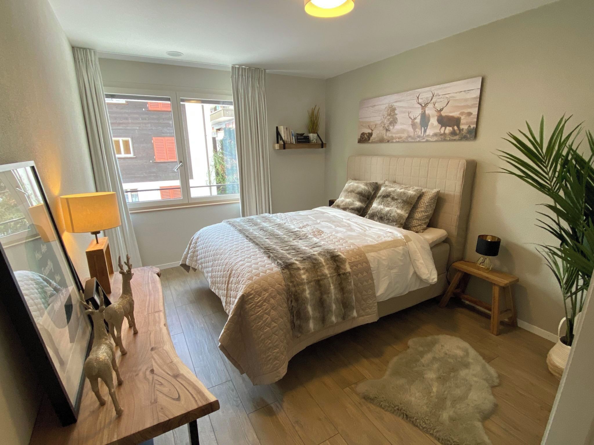 Quarto com cama, mesas de cabeceira, luminária, espelho, janela com cortinas, prateleira na parede e um quadro.