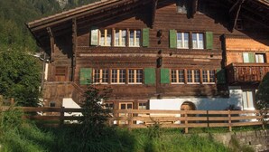 En traditionel træchalet med mange vinduer og et skråt tag, omgivet af grøn bevoksning og et bjergrigt landskab.