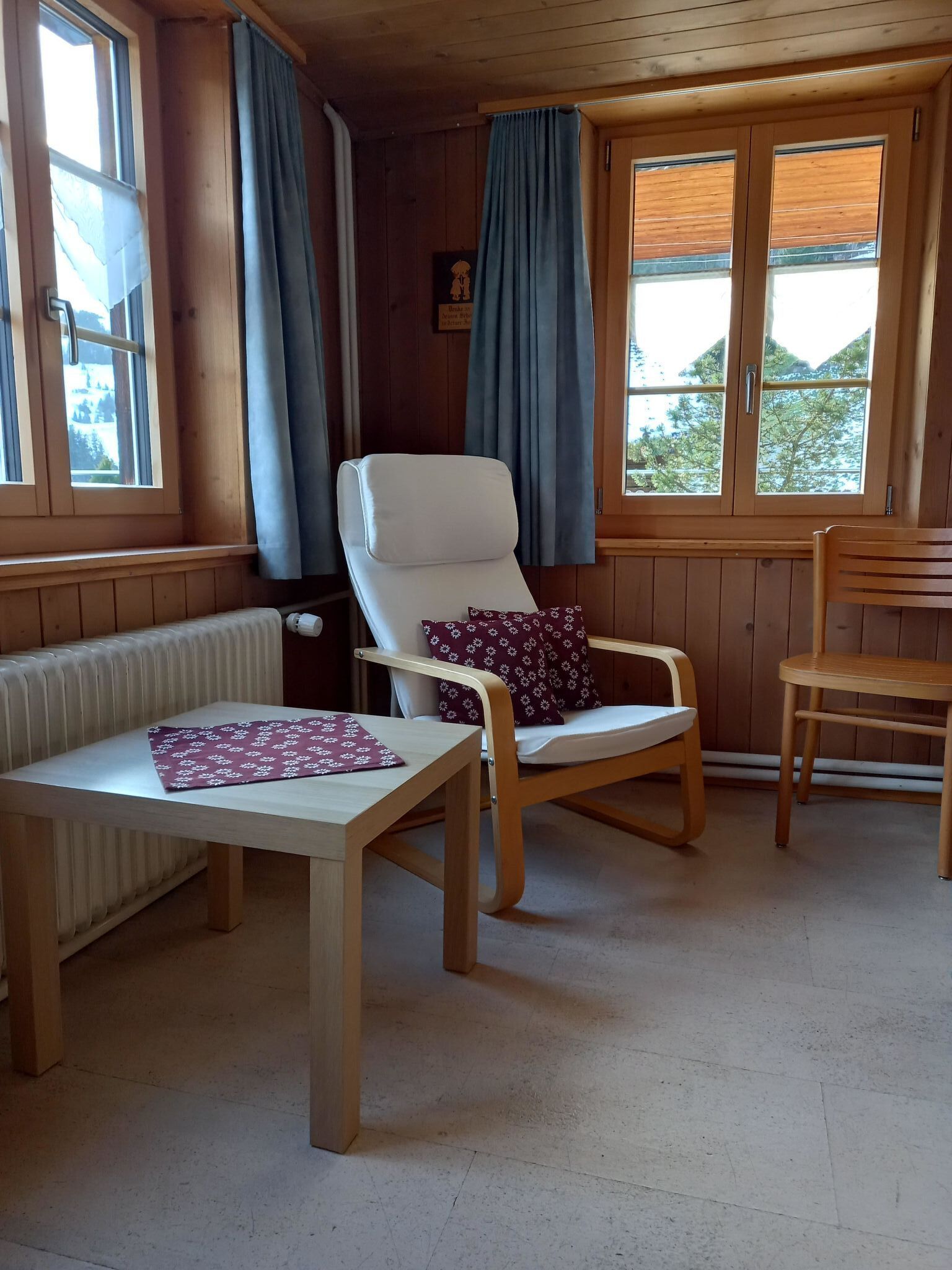 A room with a wooden chair, a table, and a bench. There are windows with curtains, a radiator, and a wooden door.