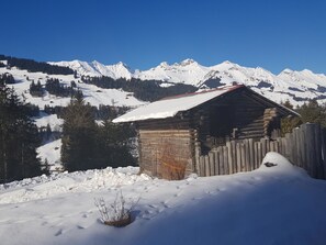 Eine Holzhütte mit schneebedecktem Dach, umgeben von einem Holzzaun, vor einer Kulisse aus schneebedeckten Bergen.