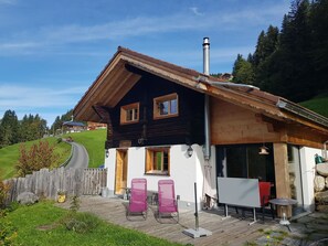 A two-story wooden house with a sloped roof, a chimney, and a small outdoor seating area with chairs. - Chalet Giisställi Ferienhaus, Dusche, WC Ferienhaus by Interhome (Adelboden)