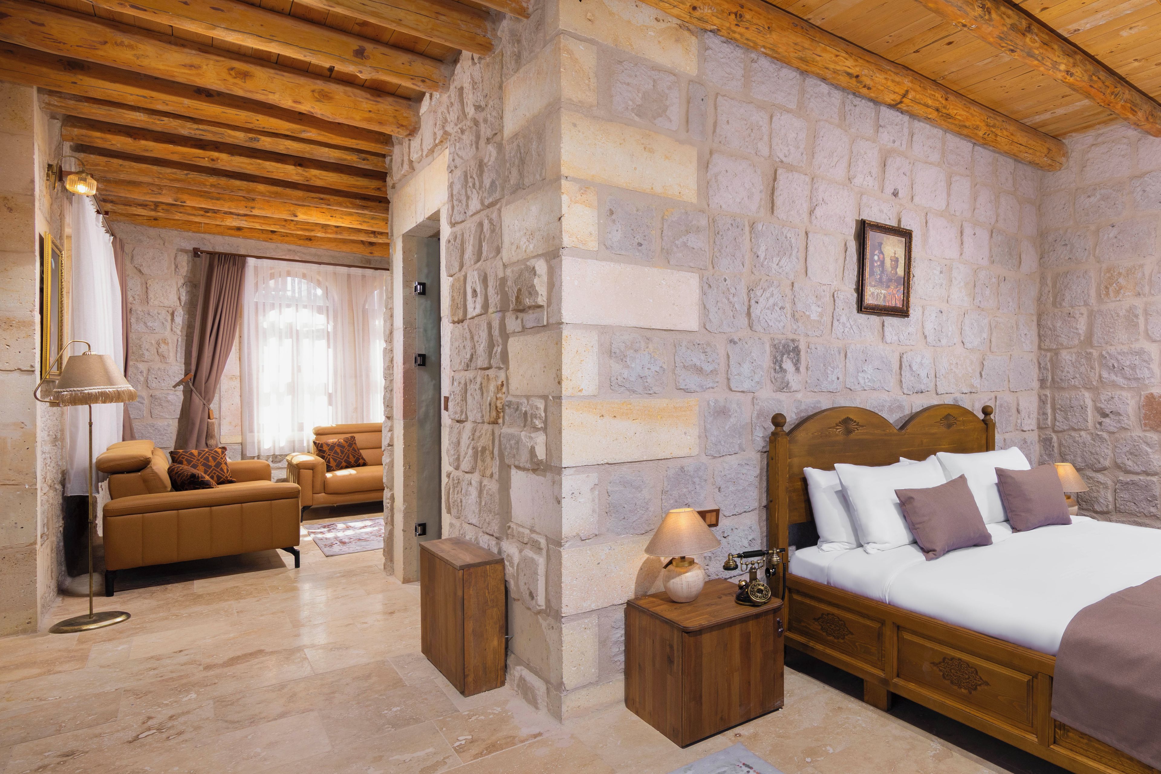 Un dormitorio con paredes de piedra, techo de madera, cama con ropa blanca, mesita de noche y sofá con cojines estampados.