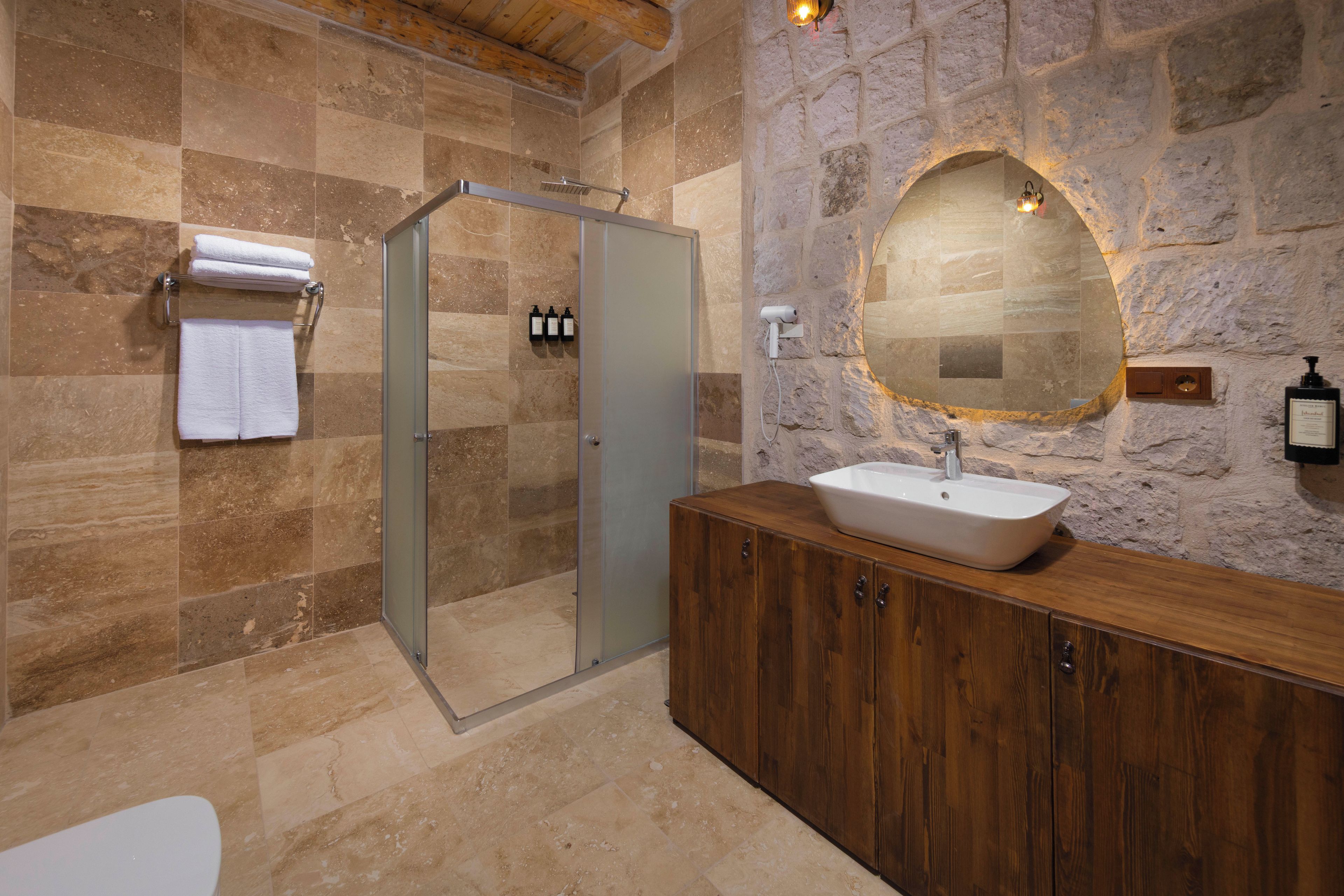 Un baño con cabina de ducha de vidrio, tocador de madera con lavamanos y pared de piedra con un espejo grande.