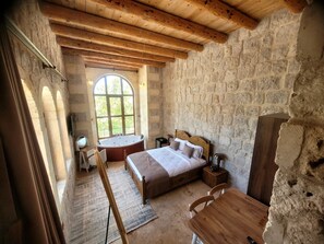 A rustic bedroom with a stone wall, a wooden ceiling, a large arched window, a wooden bed, a bedside table, and a wooden chair.