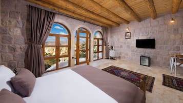 A room with a large bed, stone walls, wooden beams, a TV, and a view through arched windows.