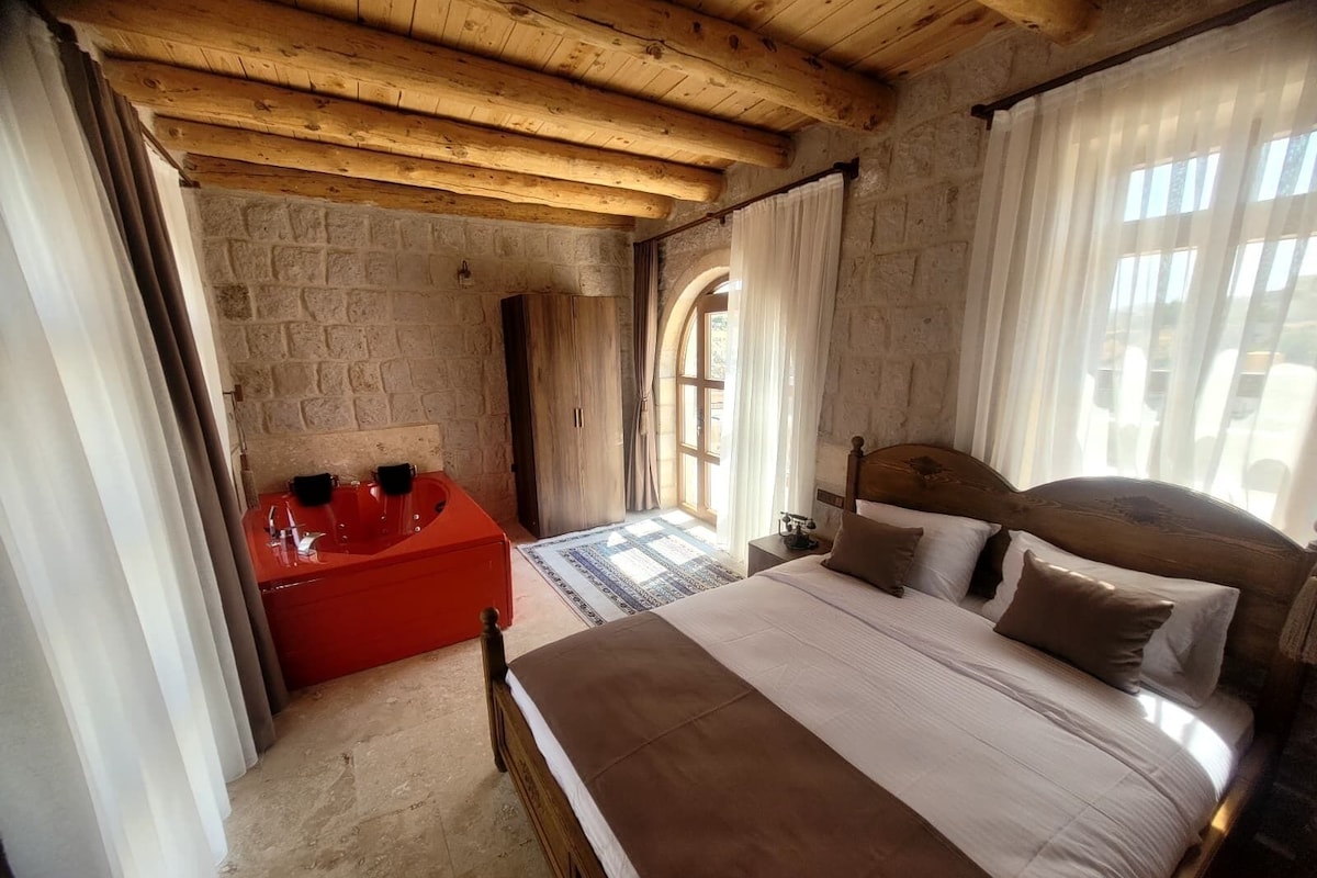A bedroom with a wooden ceiling, stone walls, a large bed with pillows, a red bathtub, and a window with curtains.