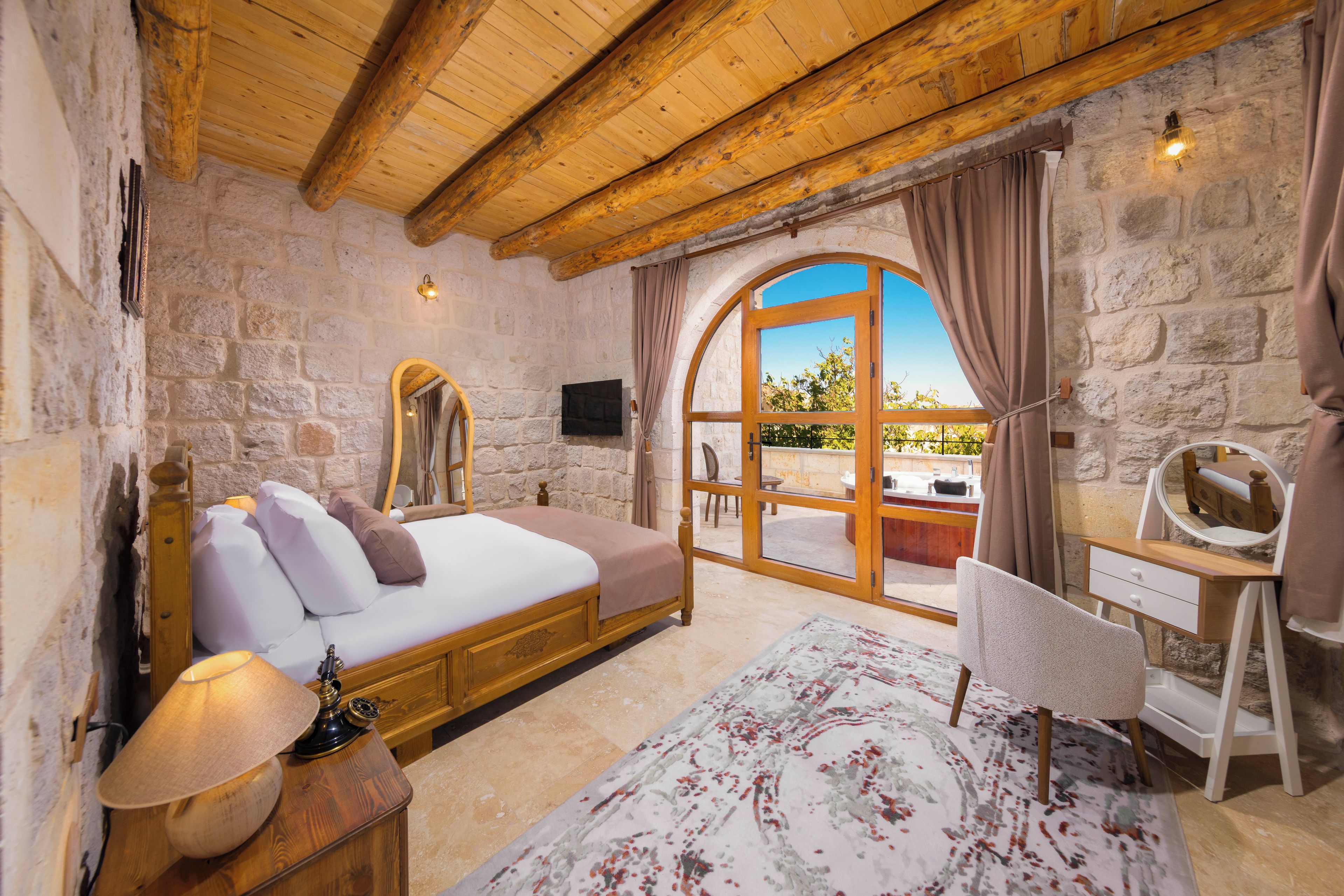 Un dormitorio con pared de piedra, vigas de madera en el techo, una cama, un escritorio, una silla y un amplio ventanal de arco.