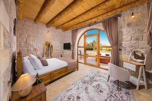 A bedroom with a stone wall, wooden ceiling beams, a bed, a desk, a chair, and a large arched window.