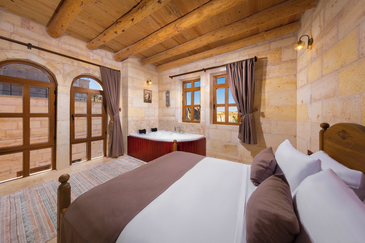 A room with a large bed, a wooden ceiling, stone walls, a window, and a bathroom with a bathtub.